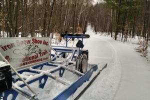 A picture of a snow packed road leading through trees, with an Iron Snow Shoe Inc. Recreational Trail sign in front.