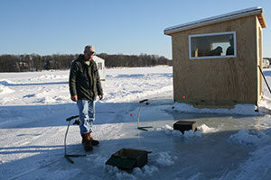 An image of a man ice fishing in front of a shack.